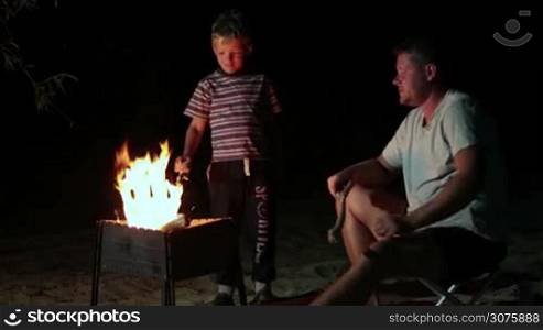 Family have a rest in their camp at night near campfire. Son casting log in burning fire.