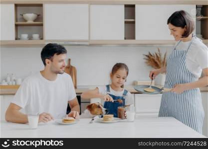 Family have breakfast at kitchen in morning. Happy girl puts melted chocolate on tasty fried pancake, poses at table with father, dog, mother stands near, wears apron, holds pan, busy cooking