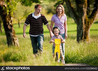 Family having a walk outdoors in summer, their little son using a training bike, unspoiled nature
