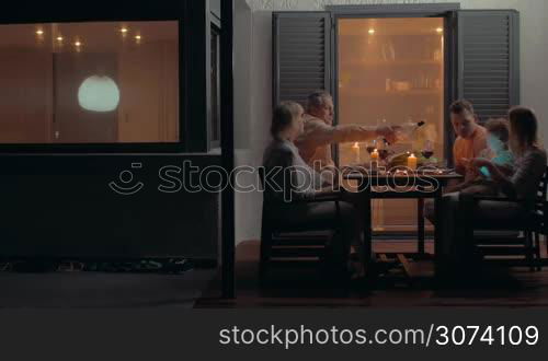 Family having dinner outdoor in backyard. Parents with son and grandparents enjoying summer evening and tasty meal. Boy playing on pad with mother