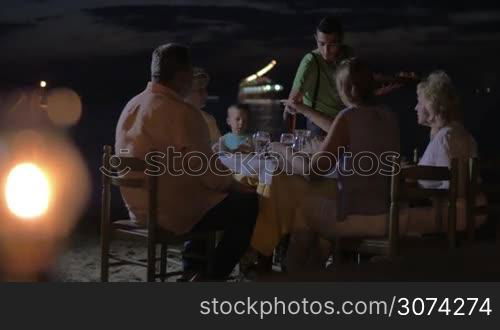 Family having meal in outdoor restaurant on the beach at night. Waiter serving dinner, burning tiki torch in foreground