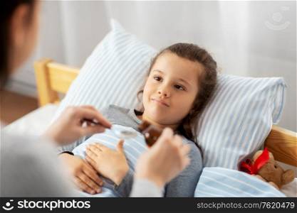 family, health and medicine concept - mother pouring cough syrup for little sick daughter lying in bed at home. mother pouring cough syrup for sick daughter
