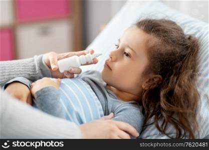 family, health and medicine concept - mother with nasal spray treats little sick daughter lying in bed at home. mother with nasal spray treats sick daughter