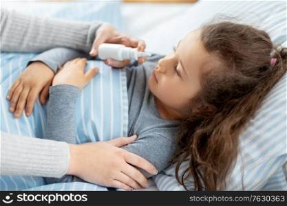 family, health and medicine concept - mother with nasal spray treats little sick daughter lying in bed at home. mother with nasal spray treats sick daughter