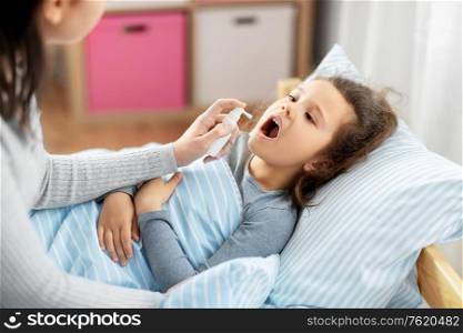 family, health and medicine concept - mother with oral spray treats little sick daughter lying in bed at home. mother with oral spray treats sick little daughter