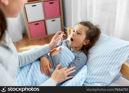 family, health and medicine concept - mother with oral spray treats little sick daughter lying in bed at home. mother with oral spray treats sick little daughter