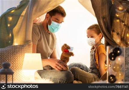 family, health and people concept - happy father with teddy bear toy and little daughter wearing face protective medical mask for protection from virus disease playing in kids tent at night at home. happy family playing with toy in kids tent at home