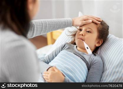 family, health and people concept - mother with sick little daughter lying in bed with oral thermometer and measuring temperature at home. mother and sick daughter measuring temperature