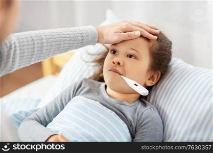 family, health and people concept - mother with sick little daughter lying in bed with oral thermometer and measuring temperature at home. mother and sick daughter measuring temperature