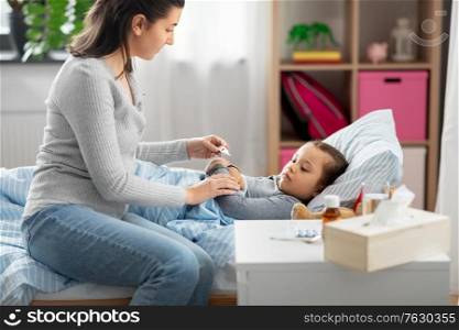 family, health and people concept - mother with thermometer measuring temperature of sick daughter lying in bed at home. mother measuring temperature of sick daughter