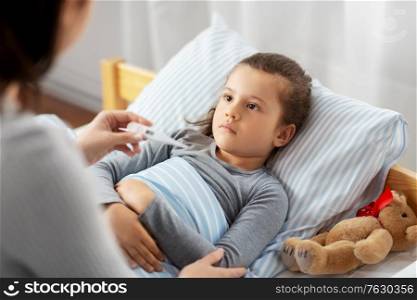 family, health and people concept - mother with thermometer measuring temperature of sick daughter lying in bed at home. mother measuring temperature of sick daughter