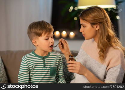 family, health and people concept - sad mother giving antipyretic medication or cough syrup to ill little son at home. mother giving medication or cough syrup to ill son