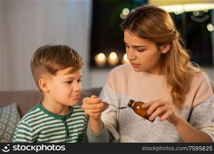 family, health and people concept - sad mother pouring antipyretic medication or cough syrup to spoon for ill little son at home. mother giving medication or cough syrup to ill son