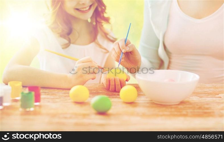 family, holiday, handcraft, people and childhood concept - close up of happy girl and mother with brushes coloring easter eggs