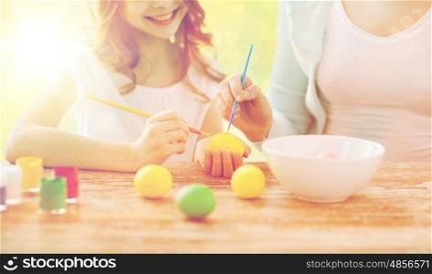family, holiday, handcraft, people and childhood concept - close up of happy girl and mother with brushes coloring easter eggs