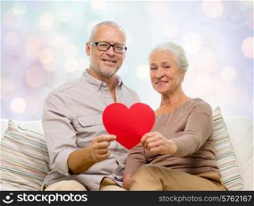 family, holidays, age and people concept - happy senior couple holding little red paper heart shape cutout and sitting on sofa over blue lights background