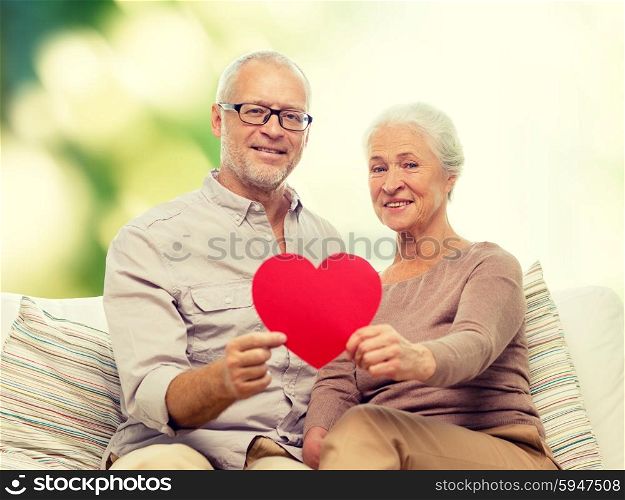 family, holidays, age and people concept - happy senior couple holding little red paper heart shape cutout and sitting on sofa over green background