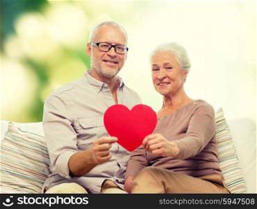 family, holidays, age and people concept - happy senior couple holding little red paper heart shape cutout and sitting on sofa over green background