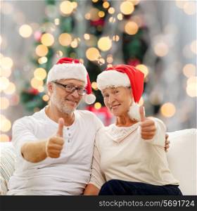 family, holidays, age and people concept - happy senior couple in santa helper hats sitting on sofa over christmas tree lights background