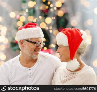 family, holidays, age and people concept - happy senior couple in santa helper hats sitting on sofa over christmas tree lights background