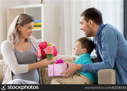 family, holidays and greetings concept - father and little son giving flowers and present to happy pregnant mother at home. family giving present to pregnant mother at home