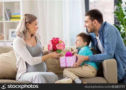 family, holidays and greetings concept - father and little son giving flowers and present to happy pregnant mother at home. family giving present to pregnant mother at home