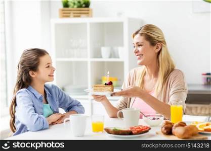 family, holidays and people concept - happy girl and mother holding piece of birthday cake with burning candle at home kitchen. happy family with birthday cake at home kitchen