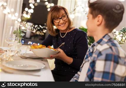 family, holidays and people concept - happy grandmother offering grandson potato at home dinner party. grandmother and grandson having dinner at home