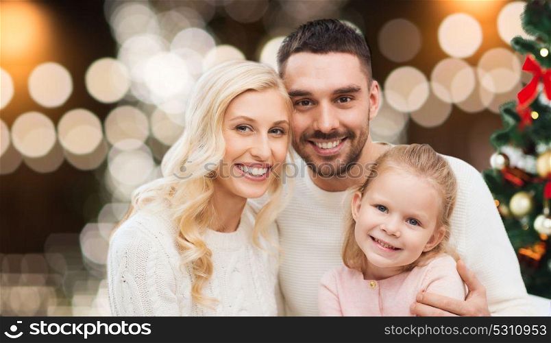 family, holidays and people concept - happy mother, father and little daughter sitting on sofa at christmas tree over lights background. happy family at home with christmas tree
