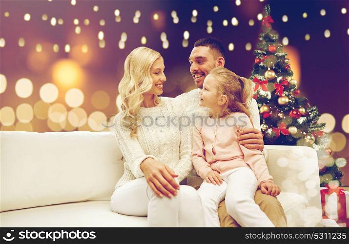 family, holidays and people concept - happy mother, father and little daughter sitting on sofa at christmas tree over lights background. happy family at home with christmas tree