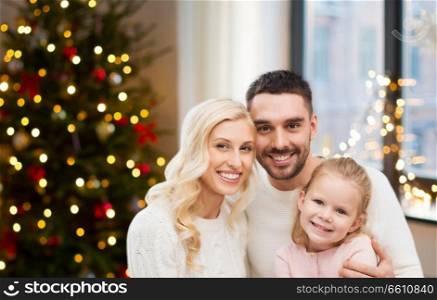 family, holidays and people concept - happy mother, father and little daughter sitting on sofa at home over christmas tree lights background. happy family at home over christmas tree lights