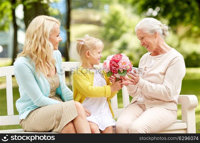 family, holidays and people concept - happy smiling granddaughter giving flowers to her grandmother sitting on park bench. happy family giving flowers to grandmother at park