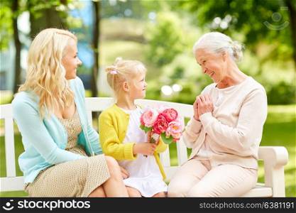 family, holidays and people concept - happy smiling granddaughter giving flowers to her grandmother sitting on park bench. happy family giving flowers to grandmother at park