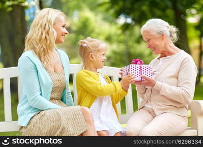 family, holidays and people concept - happy smiling granddaughter giving present to her grandmother sitting on park bench. happy family giving present to grandmother at park