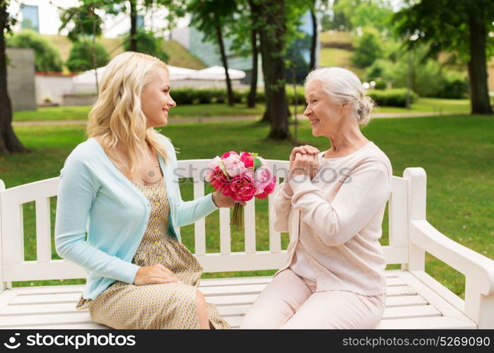 family, holidays and people concept - happy smiling young daughter giving flowers to her senior mother sitting on park bench. daughter giving flowers to senior mother at park