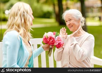 family, holidays and people concept - happy smiling young daughter giving flowers to her senior mother sitting on park bench. daughter giving flowers to senior mother at park