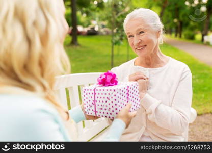 family, holidays and people concept - happy smiling young daughter giving present to her senior mother sitting on park bench. daughter giving present to senior mother at park