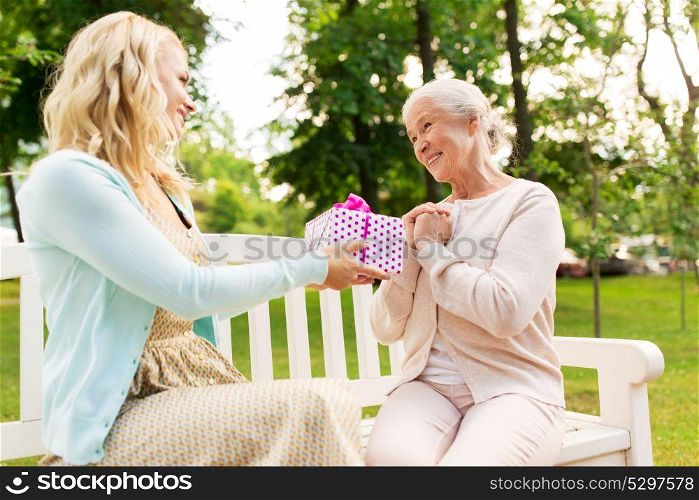 family, holidays and people concept - happy smiling young daughter giving present to her senior mother sitting on park bench. daughter giving present to senior mother at park