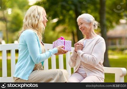 family, holidays and people concept - happy smiling young daughter giving present to her senior mother sitting on park bench. daughter giving present to senior mother at park