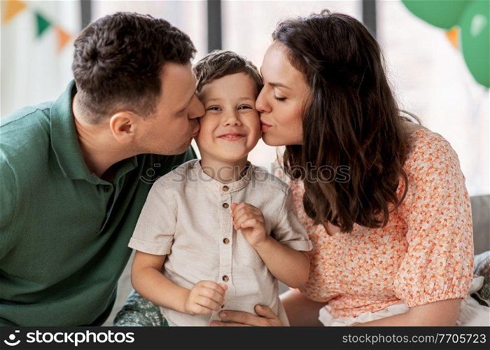 family, holidays and people concept - portrait of happy mother and father kissing little son sitting on sofa at home party. happy parents kissing little son at birthday party