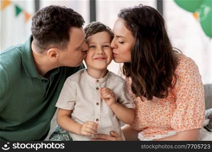 family, holidays and people concept - portrait of happy mother and father kissing little son sitting on sofa at home party. happy parents kissing little son at birthday party