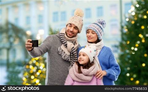 family, holidays and technology concept - happy mother, father and little daughter in winter clothes taking selfie by smartphone over christmas tree lights on city street background background. happy family taking selfie by smartphone