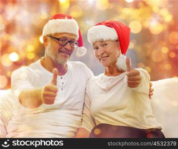 family, holidays, christmas, age and people concept - happy senior couple in santa helper hats sitting on sofa and showing thumbs up gesture over lights background