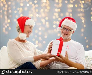 family, holidays, christmas, age and people concept - happy senior couple in santa helper hats with gift box over lights background
