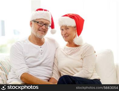 family, holidays, christmas, age and people concept - happy senior couple in santa helper hats sitting on sofa at home