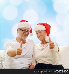 family, holidays, christmas, age and people concept - happy senior couple in santa helper hats sitting on sofa over blue lights background and showing thumbs up gesture