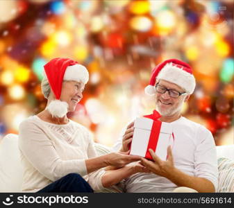 family, holidays, christmas, age and people concept - happy senior couple in santa helper hats with gift box over red lights background
