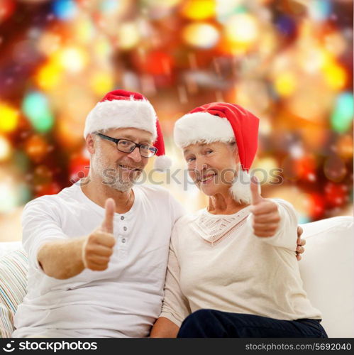 family, holidays, christmas, age and people concept - happy senior couple in santa helper hats sitting on sofa over red lights background and showing thumbs up gesture