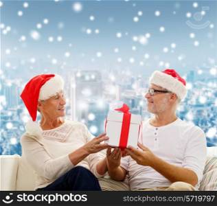 family, holidays, christmas, age and people concept - happy senior couple in santa helper hats with gift box over snowy city background
