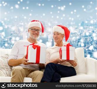 family, holidays, christmas, age and people concept - happy senior couple in santa helper hats with gift boxes over snowy city background